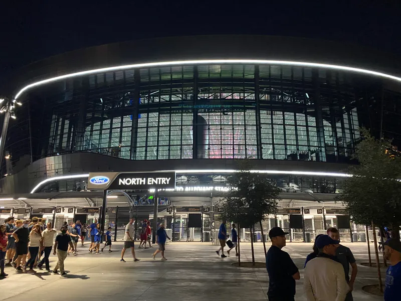 The glass north entry of Allegiant Stadium lit at night with fans walking toward the gates