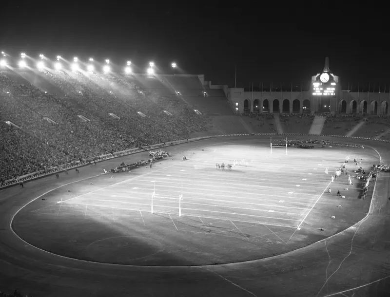 Night view of a packed Los Angeles Memorial Coliseum under floodlights, the peristyle and scoreboard at the far end