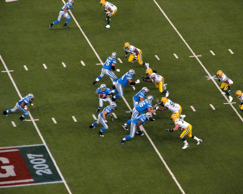 Elevated view of the Lions running a handoff against the Packers on the Ford Field turf, the Thanksgiving 2007 logo painted near the sideline